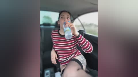 Media: Video of a young woman with dark hair, wearing a striped sweater and shorts, drinking water from a bottle inside a car, blurred background.