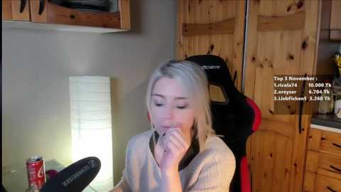 Media: Video of a blonde woman in a beige sweater, drinking from a can, sitting at a wooden desk with a lamp and red and black gaming chair.