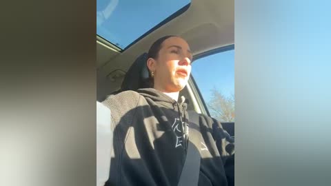 Media: A video of a woman with fair skin, dark hair, wearing a black hoodie, and a white shirt, driving a car with a clear blue sky visible through the window.