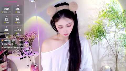 Media: Video of an Asian woman with long black hair, wearing a white off-shoulder top, and bear headband, in a softly lit room with a potted plant.