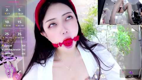 Media: Video of a young Asian woman with fair skin, wearing a red beret and white robe, gagged with a red ball gag. Background shows a blurred, green-lit room with plants.