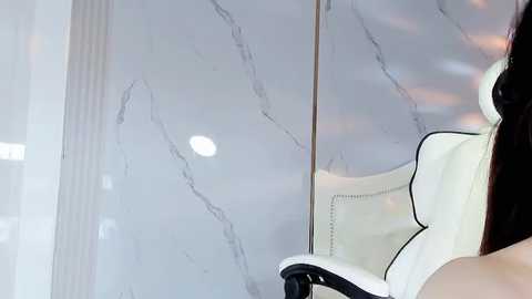 Media: A video of a sleek, modern white leather chair with black armrests in a minimalist room with large, marbled glass walls reflecting soft light and a blurred, warm-toned background.