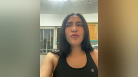 Media: Video of a young woman with medium skin tone and straight black hair, wearing a black sports bra, standing in a kitchen with white cabinets and a window.