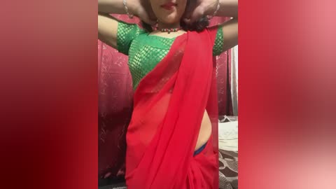 Media: Video of a South Asian woman with medium skin tone, wearing a green lace top and a red sari, hands raised, red curtains in the background.