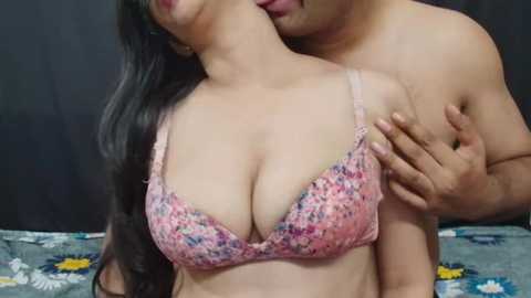 Media: A video of a topless man embracing a woman in a floral bra, kissing her neck. She has long black hair, light skin, and large breasts. Background is a plain black wall.