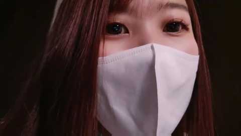 Media: Video of an Asian woman with long, straight brown hair and light skin, wearing a white surgical mask, eyes visible behind the mask.