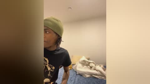 Media: Video of a young Black woman with braided hair, wearing a green beanie and black t-shirt with a graphic design, standing in a cluttered bedroom with beige walls, a blue bedspread, and a messy room.