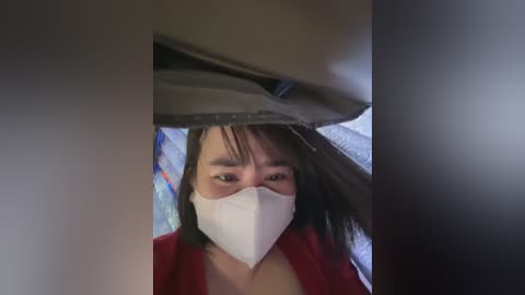 Media: Video of a woman with short, dark hair and a white surgical mask, wearing a red jacket, sitting in a car with a partially open sunroof.