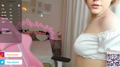 Media: A video of a young woman in a white off-the-shoulder top, standing beside a pink neon-lit bedroom with a cozy bed and plush toys.