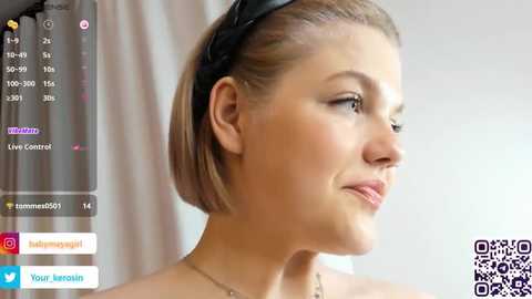 Media: Video of a blonde woman with a short bob haircut, wearing a black headband, looking to the side with a neutral expression. Background shows a live stream interface with social media icons.