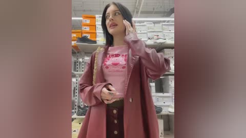 Media: Video of a young woman with long black hair, wearing a maroon coat over a matching sweater and skirt, holding a phone. She stands in a brightly lit shoe store with shelves of shoes and boxes in the background.