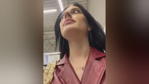 Media: A video of a woman with long black hair, wearing a red leather jacket and a necklace with an orange pendant, looking upward in a dimly lit room with exposed ceiling tiles.