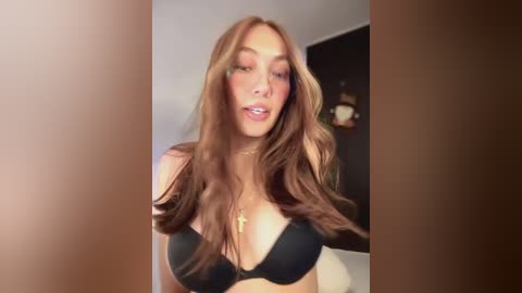 Media: Video of a young woman with long, wavy brown hair, wearing a black bra, standing in a dimly lit room with a black wall and a framed picture.