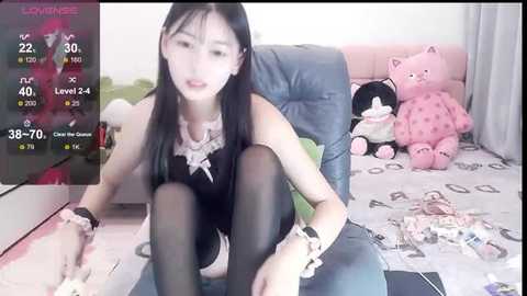 Media: A video of an Asian woman with long black hair, wearing a black lace bra and pantyhose, sitting on a couch surrounded by stuffed toys, playing a video game.