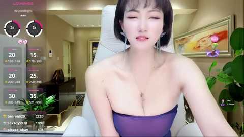 Media: A video of a young East Asian woman with short black hair, wearing a sheer purple strapless top, sitting in a modern living room.