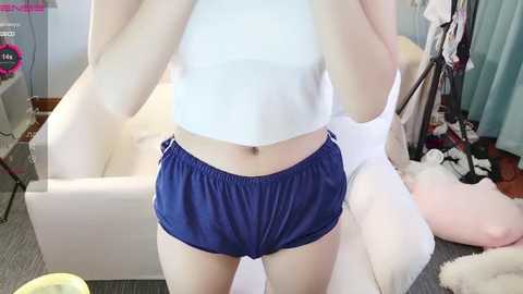 Media: Video of a young woman wearing a white crop top and blue shorts, standing in a messy bedroom with a white bed and clothes scattered on the floor.