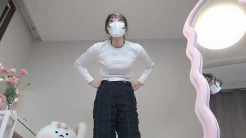 Media: Video of a woman with light skin, wearing a white mask, long-sleeve white shirt, high-waisted black pants, standing confidently with hands on hips in a minimalistic room with a round mirror and floral decorations.