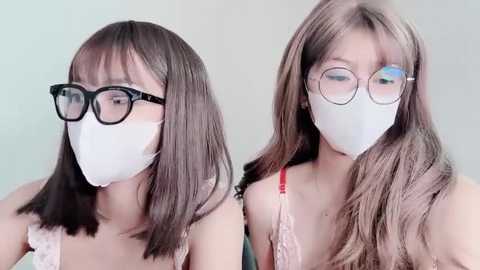 Media: Video of two young women with long brown hair, wearing white face masks, black-framed glasses, and light-colored tops, against a light-colored background.