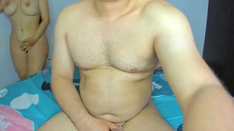 Video of a naked, overweight man with a large belly, light skin, and small penis. Background shows a naked woman standing, light skin, small breasts, and partially visible face. Both are on a bed with blue and white patterned sheets.