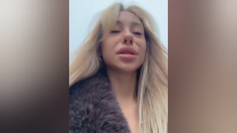 Media: Video of a young woman with long, blonde hair, wearing a dark fur coat, looking down with a neutral expression against a blurred, out-of-focus background.