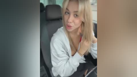 Media: Video of a blonde woman with fair skin, wearing a white shirt, sitting in a car, with blurred background.