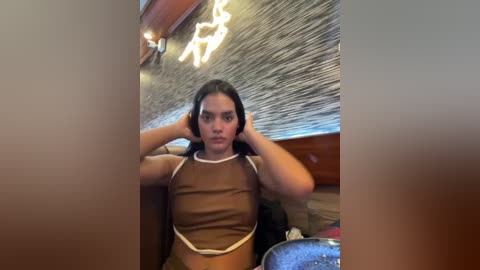 Media: Video of a young woman with long dark hair, fair skin, and a medium build, wearing a brown crop top, adjusting her headphones, against a wooden and textured wall background.