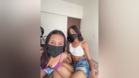 Media: Video of two young women, one with light brown skin and long hair, the other with dark skin and short hair, wearing face masks and casual clothing, sitting on a couch in a simple room.