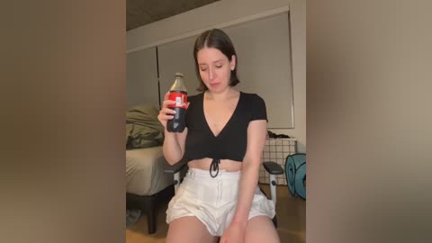 Video of a young woman with light skin and short brown hair, wearing a black crop top and white shorts, holding a red Coke bottle, in a dimly lit bedroom with a bed, clothes, and a blue backpack in the background.