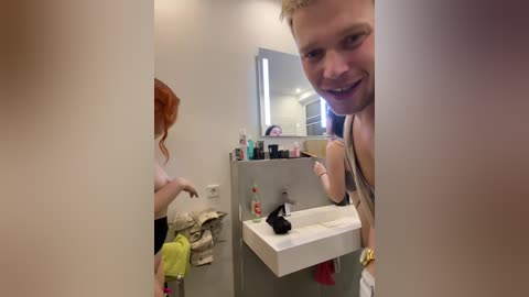 Media: A video of a bathroom with a young man with short blond hair, smiling, peering over a white sink. The sink is cluttered with toiletries and a towel. A woman with red hair, wearing a white top, is on the left, washing her hands.