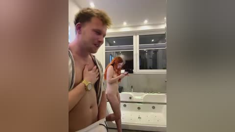 Media: Video of a young man with fair skin and short blonde hair, shirtless, holding a phone, standing in a modern, dimly lit bathroom with a large window and a white bathtub.