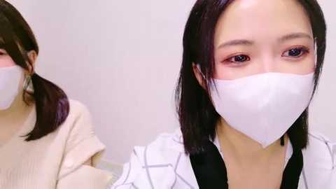 Media: Video of an Asian woman with straight black hair, wearing a white surgical mask and a white and black checkered lab coat, sitting in a sterile, white-lit room.