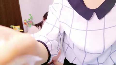 Media: Video of a woman in a white, grid-patterned blouse with a dark collar, bending over to pet a dog. Background shows a blurred indoor setting with flowers and a door.