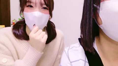 Media: Video of a young Asian woman with long black hair, wearing a white face mask, beige sweater, and a white shirt, holding a finger to her lips, sitting on a bed.