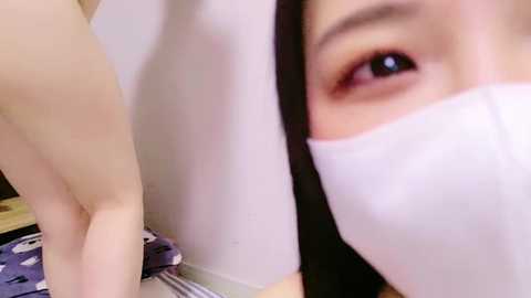 Media: Video of an Asian woman with pale skin, wearing a white face mask, looking directly at the camera, revealing her brown eyes. Background includes a white wall and a purple-patterned fabric.