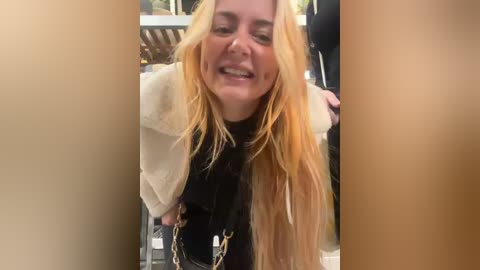 Media: Video of a blonde woman with long, wavy hair, wearing a white fur coat and black accessories, smiling joyfully outdoors.