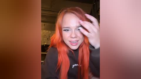 Media: Video of a young woman with long, vibrant orange hair, smiling, touching her face, in a dimly lit room with a kitchen background.