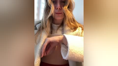 Media: Video of a blonde woman in a cozy, cream-colored fleece jacket, adjusting her bra. She has fair skin, long wavy hair, and a neutral expression. Soft lighting, blurred background.