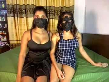 Media: Video of two women wearing black face masks, black tank tops, and black skirts on a green bed, in a bedroom with yellow curtains and a bookshelf.