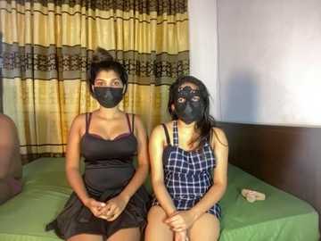 Media: Video of two women with dark skin, wearing black masks and revealing lingerie, sitting on a bed with green sheets, in a dimly lit room with yellow curtains.