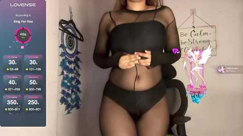 Media: Video of a woman in black mesh top and panties, holding a smartphone, standing in a room with dreamcatchers, \"Be Calm and Be Cheerful\" sign, and a chair.