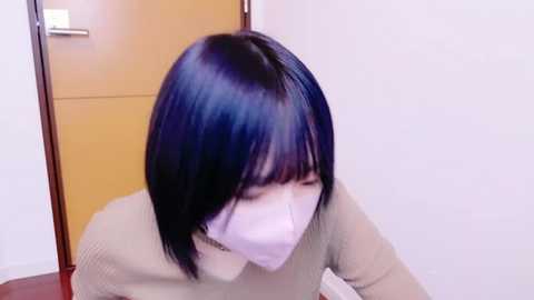 Media: Video of an Asian woman with shoulder-length black hair, wearing a beige sweater and a white mask, kneeling on a wooden floor in a hallway with a brown door and white walls.