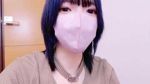 Media: Video of a young person with straight, shoulder-length blue hair, wearing a light pink surgical mask and a beige ribbed sweater, with a metal chain necklace. Background shows a wooden door and white wall.