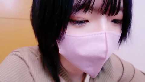 Media: Video of an East Asian woman with straight black hair, wearing a light pink surgical mask and beige sweater, focusing intently on something off-frame.