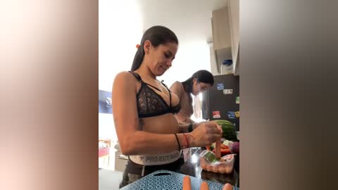 Media: Video of a fit, dark-haired woman in a black lace bra and grey shorts, slicing vegetables on a countertop, with another woman in the background.