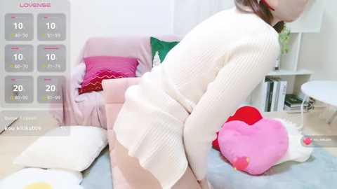 Media: Video of a young woman in a white, textured cardigan, leaning over a pink sofa with colorful pillows, surrounded by playful, heart-shaped plush toys. The room has a modern, minimalist decor.