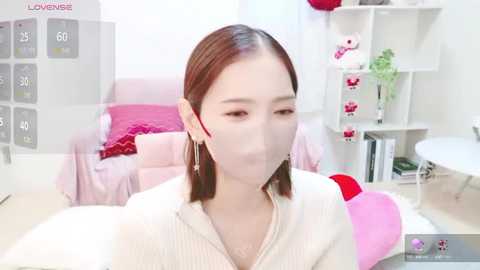 Media: Video of an East Asian woman with fair skin and straight brown hair, wearing a white ribbed sweater, a face mask, and pink headphones, sitting in a modern, minimalist bedroom with pink furniture, a white shelf, and a small plant.