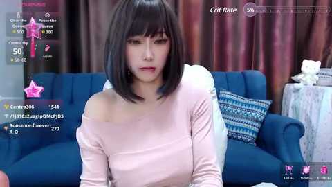 Media: Video of an East Asian woman with a pale complexion and short black bob haircut, wearing a light pink off-shoulder top, seated in a blue sofa with patterned pillows, indoors with brown curtains.