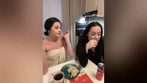 Media: A video captures two young women, one with fair skin and dark hair, wearing a beige towel, and another with olive skin and long black hair, dressed in black, eating cereal in a modern kitchen.