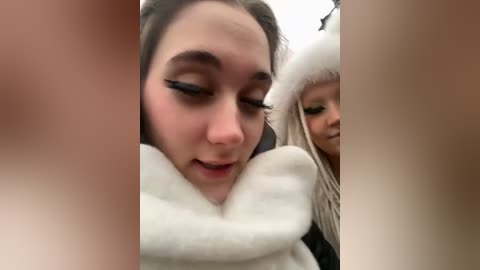 Media: A blurry video of a young woman with dark hair, wearing thick black eyeliner and a white furry coat, sitting next to a child with blonde hair and a white hat.