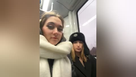 Media: Video of two women in a bus: one with light skin, dark hair, and a white scarf, the other with pale skin, blonde hair, and a black hat.